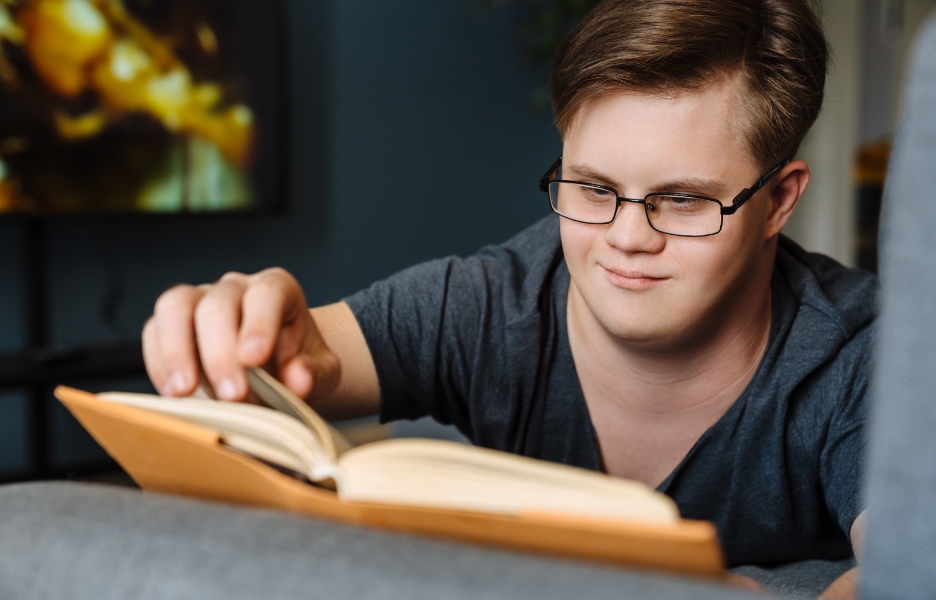 A special needs boy with glasses is reading a book and smiling about what he has just read.