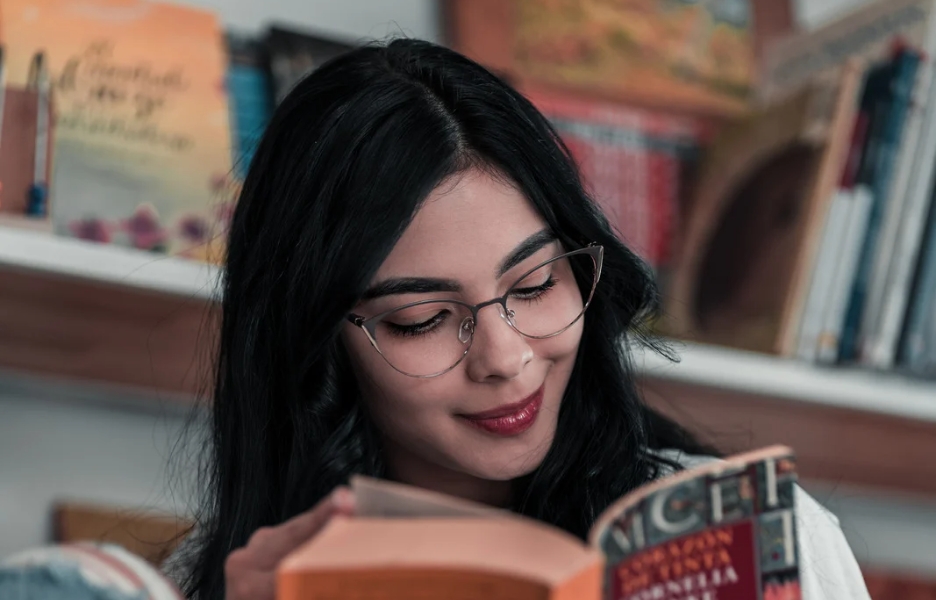 A woman with black hair and glasses smiles at the book that she is reading.